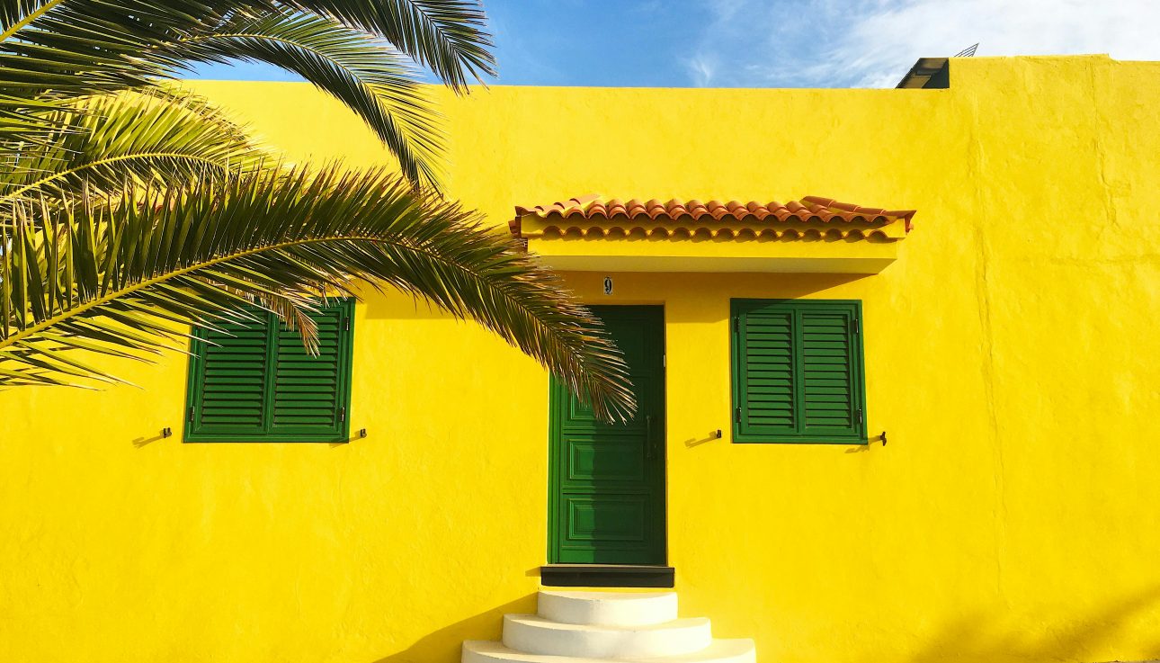 Vibrant yellow house facade in Spain with green shutters and a palm tree.